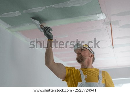 The worker make a plasterboard ceiling. He does taping plaster drywall ceiling joints. He is using special stainless steel taping knife.