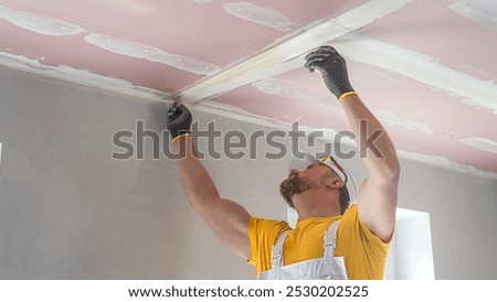 The worker make a plasterboard ceiling. He does taping plaster drywall ceiling joints. He is inserting the glass tape into the putty.
