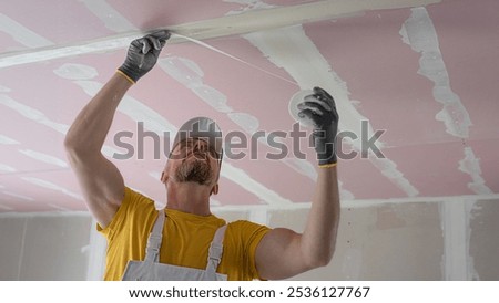 The worker make a plasterboard ceiling. He does taping plaster drywall ceiling joints. He is  putting fiberglass tape into the plaster putty. 