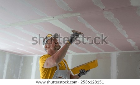 The worker make a plasterboard ceiling. He does taping plaster drywall ceiling joints. He is using special drywall taping knife.
