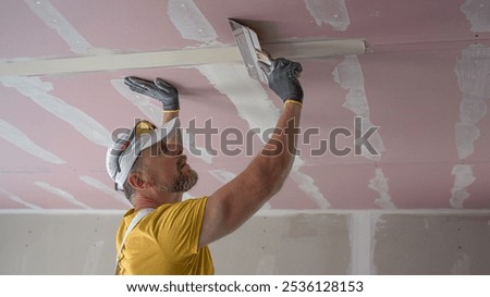The worker make a plasterboard ceiling. He does taping plaster drywall ceiling joints. He is using special drywall taping knife.