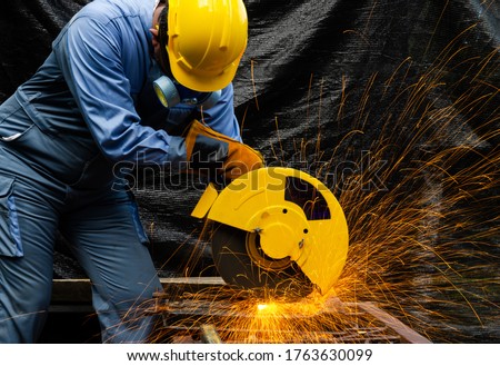 Worker man in blue uniform with yellow satety helmet and leather gloves cutting metal pipe by electric circular abrasive saw with sparks. Industrial working with personnel protective equipment concept
