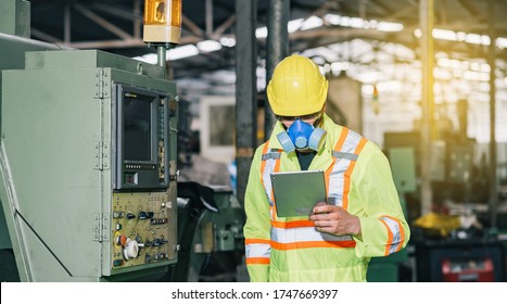 Worker Man Caucasian  Wear A Blue Protective Mask In Safety Jumpsuit Uniform With Yellow Hardhat And Using Tablet At Factory.Metal Working Industry Concept Professional Engineer Manufacturing Machine
