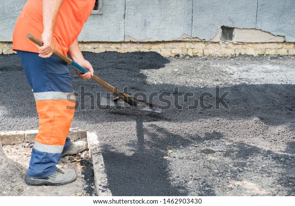 Worker\
man lays asphalt road repair road paving yellow sun ray light. A\
man in overalls is laying asphalt with a\
shovel