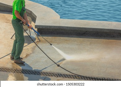 Worker Man In Uniform Washes Street Or Park Sidewalk Near Water Pool Or Fountain. Municipal Service Of City Cleaning Process. Guy Uses Spray Equipment