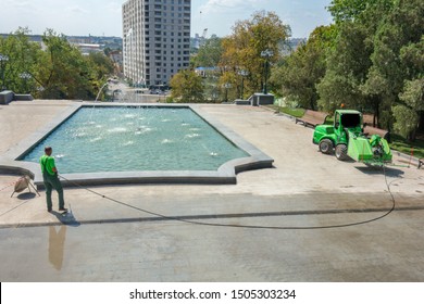 Worker Man In Uniform Washes Street Or Park Sidewalk Near Water Pool Or Fountain. Municipal Service Of City Cleaning Process. Guy Uses Spray Equipment Machine