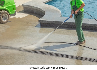 Worker Man In Uniform Washes Street Or Park Sidewalk Near Water Pool Or Fountain. Municipal Service Of City Cleaning Process. Guy Uses Spray Equipment Machine