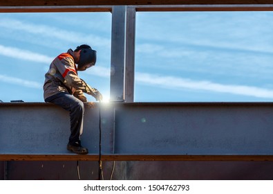 Worker Man welder in uniform of a welder sitting on an iron support welds an iron beam against the sky - Powered by Shutterstock