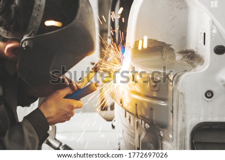 Worker in mask welds car in auto service of bodywork.