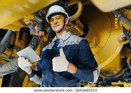 Worker mechanic with tablet computer under mining truck in garage, service center of industrial machinery.