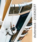 A worker in mountaineering equipment paints the hull of a white ship with a paint roller at a height