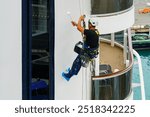 A worker in mountaineering equipment paints the hull of a white ship with a paint roller at a height