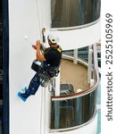 A worker in mountaineering equipment paints the hull of a white ship with a paint roller while hanging from a rope