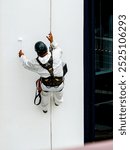 A worker in mountaineering equipment paints the hull of a white ship with a paint roller while hanging from a rope