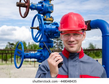 Worker Near Well Head Valve Holding Radio And Wearing Red Helmet In The Oilfield. Oil And Gas Concept. 