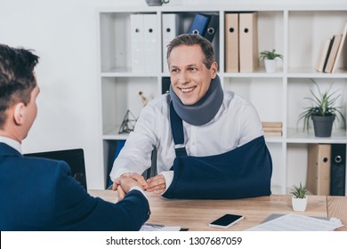 worker in neck brace with brokenarm and businessman in blue jacket shaking hands over table in office, compensation concept