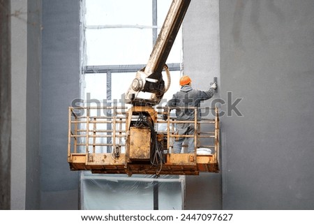 Worker on crane plastering building facade wall, construction worker in hardhat, work at height. Revamping building exteriors, stucco application by skilled worker and crane operation