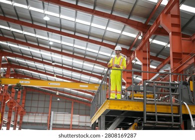 A worker is on a high platform in a warehouse. The worker wears a hard hat and a safety vest. The warehouse has high ceilings and large machinery inside.