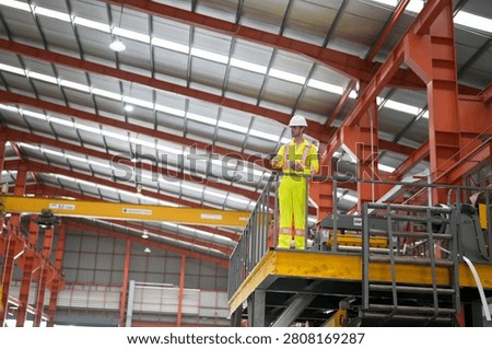 A worker is on a high platform in a warehouse. The worker wears a hard hat and a safety vest. The warehouse has high ceilings and large machinery inside.