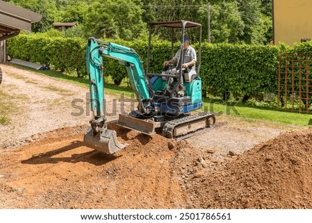 A worker on a mini excavator is digging the ground ahead of the house. Groundwork and exterior paving.