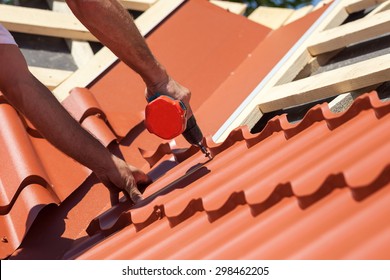 Worker on a roof with electric drill installing red metal tile on wooden house - Shutterstock ID 298462205
