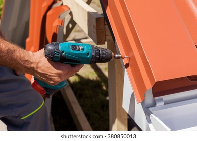 Worker On A Roof With Electric Drill Installing Red Metal Tile On Wooden House