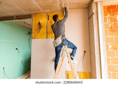 Worker on wooden ladder holds professional manual power tool ACU drill and tightens the screws on plasterboards installed on the partition dry wall insulated with mineral wool.