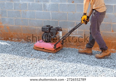 Worker operates vibration plate compactor on gravel ground next to concrete slab at construction site