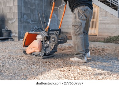 Worker operates a vibratory plate compactor on a gravel surface at a construction site. The machine compacts the ground to create a stable base for paving or structural work.