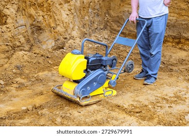 Worker operates yellow compactor machine on dirt construction site worker on leveling ground of building preparation area.