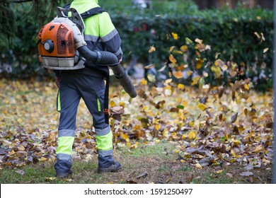Worker operating heavy duty leaf blower in city park. Dark blue and fluor green dress.Removing fallen leaves in autumn. Leaves swirling up. - Shutterstock ID 1591250497