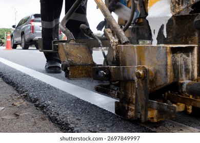 Worker operating road marking machine applying white line on new asphalt pavement. Road maintenance and construction concept, ensuring traffic safety and infrastructure quality.