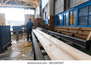 Worker operating wood processing machinery at a sawmill, shaping and rounding timber logs on production line with sawdust covering the floor, motion blur showing industrial activity in workshop.