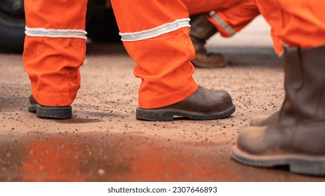 The worker in orange coverall and leather safety shoe is standing on the dirt gorund at construction worksite. Industrial worker in action scene. selective focus.