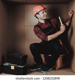 Worker In Orange Helmet Sitting In Cardboard Box. Young Man In Uniform. Uncomfortable Life. Personal Spase Concept. Uncomfortable House Concept. Young Introvert. Worker With Tools.