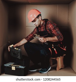 Worker In Orange Helmet Sitting In Cardboard Box. Young Man In Uniform. Uncomfortable Life. Personal Spase Concept. Uncomfortable House Concept. Young Introvert. Worker With Tools.