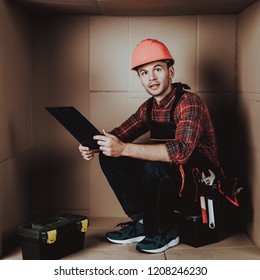 Worker In Orange Helmet Sitting In Cardboard Box. Young Man In Uniform. Uncomfortable Life. Personal Spase Concept. Uncomfortable House Concept. Young Introvert. Worker With Tools.