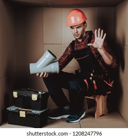 Worker In Orange Helmet Sitting In Cardboard Box. Young Man In Uniform. Uncomfortable Life. Personal Spase Concept. Uncomfortable House Concept. Young Introvert. Worker With Tools.