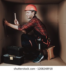 Worker In Orange Helmet Sitting In Cardboard Box. Young Man In Uniform. Uncomfortable Life. Personal Spase Concept. Uncomfortable House Concept. Young Introvert. Worker With Tools.