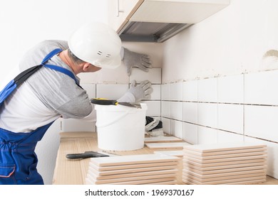 a worker in overalls applies glue to the tile with a spatula to lay it on the wall in the kitchen