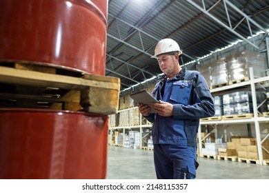 A Worker In Overalls And A Helmet In A Warehouse Inspects Products Stored In Barrels. Calculation And Verification Of Reserves In An Oil Company, Solving Logistics Problems