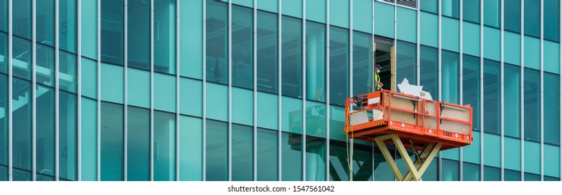 Worker In Overalls Throws Through The Window Of The Glass Business Center Debris On The Scissor Elevated Lift Platform. Banner Panorama For Header Web Site.