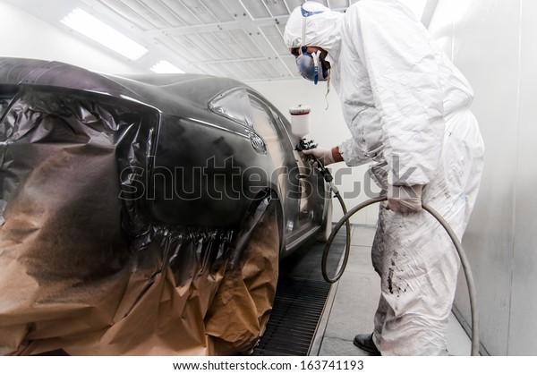 worker painting a car in a special\
painting box, wearing a white costume and protection\
gear