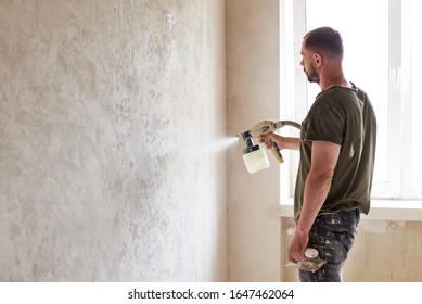 Worker Painting Wall With Spray Gun. Man With A Beard Is Dressed In Paint-smeared Jeans And A T-shirt Against The Background Of A Small Window In The Apartment. Repair Concept