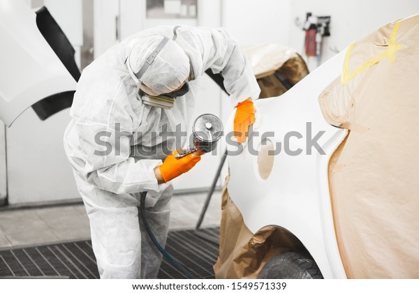 Worker paints auto with spray gun. White car\
in a paint chamber during repair\
work.