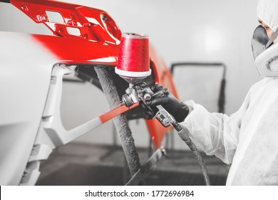 Worker Paints Bumper Of Auto With Spray Gun. Red Car Elements In A Paint Chamber During Repair Work.