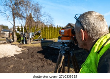 A worker peers through an orange automatic level on a tripod, surveying a yard with conifer hedge and swing set, wheelbarrow and rod holder present in bright daylight.
