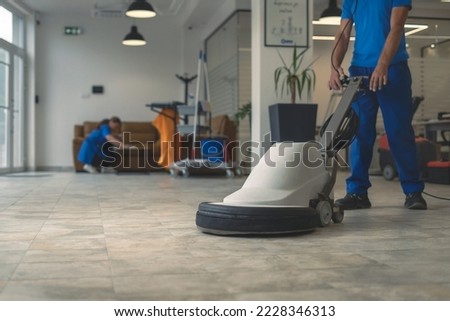 Worker polishing hard floor with high speed polishing machine while other cleaner cleans rhe table in the background