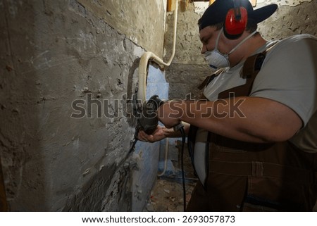 A worker prepares for a renovation, using a hammer drill to remove old plaster and cement from a wall wearing protective ear protection and a respirator mask Removing old plaster with a hammer drill.