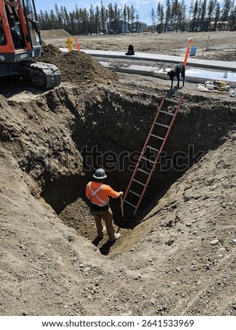 Worker Prepares Utility Excavation While Dog Waits Above
Worker levels bedding in deep utility excavation as dog waits near ladder at subdivision site.